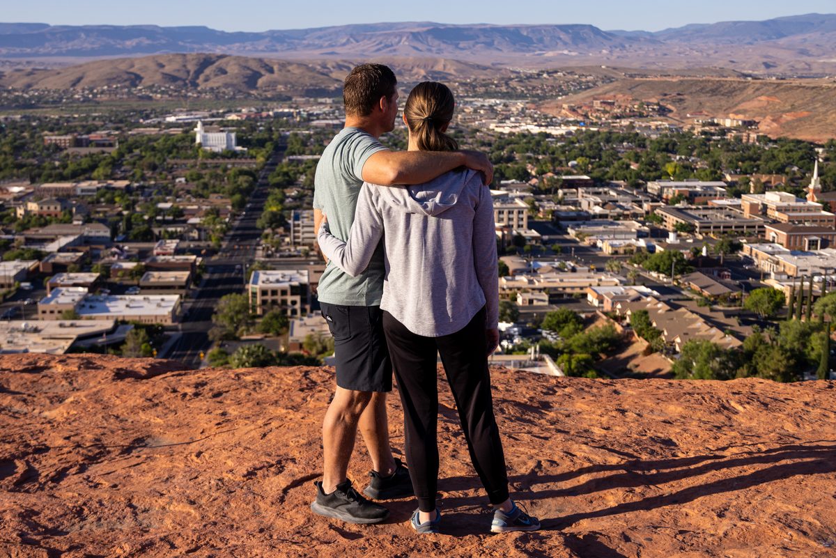 Couple stand on cliff at Pioneer Park and look down at St. George city.